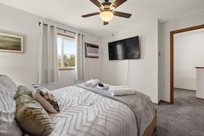 Ein Schlafzimmer mit Bett, Deckenventilator, Fernseher und einem Fenster mit Vorhängen.