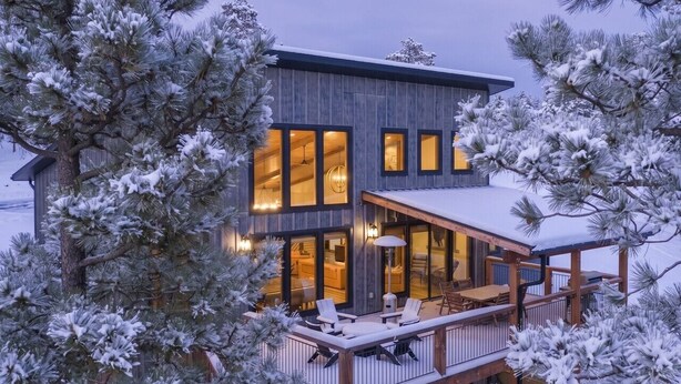 A modern house with a deck, large windows, and snow-covered trees.