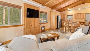 A modern cabin interior with a living area, kitchen, and dining space, featuring wooden beams, large windows, and a flat-screen TV.