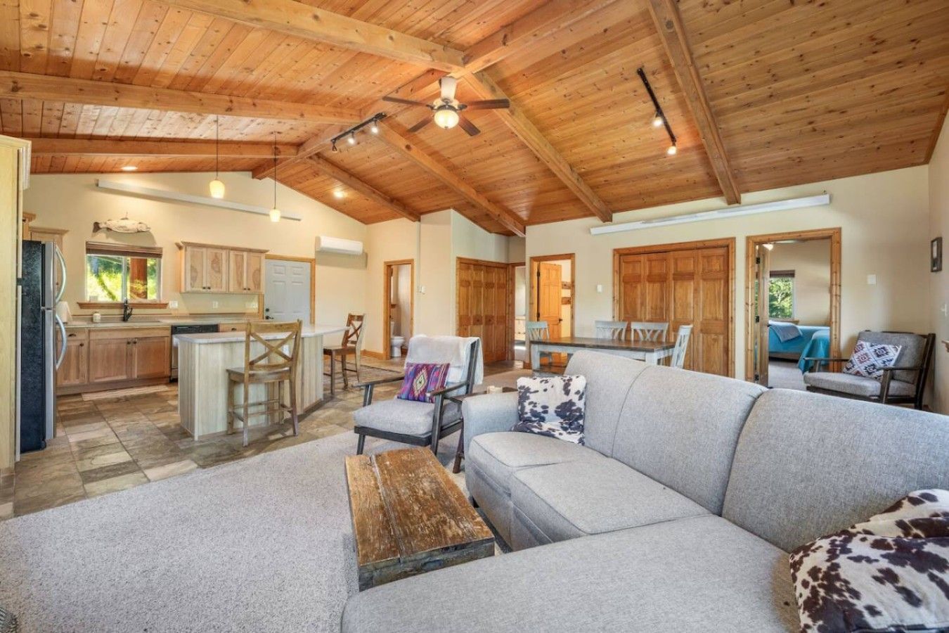 A spacious living area with a grey sofa, wooden ceiling beams, and a kitchen in the background.
