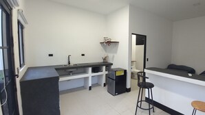 A compact kitchen with a sink, stove, and refrigerator, a small bar area with stools, and a bed with pillows. - Nature Sanctuary Studio #2 w/ AC, Close to Tamarindo, Flamingo Beaches (Tamarindo)