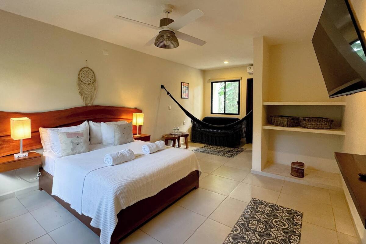 A hotel room with a large bed, a ceiling fan, a hammock, a TV, and a view of the outdoors.