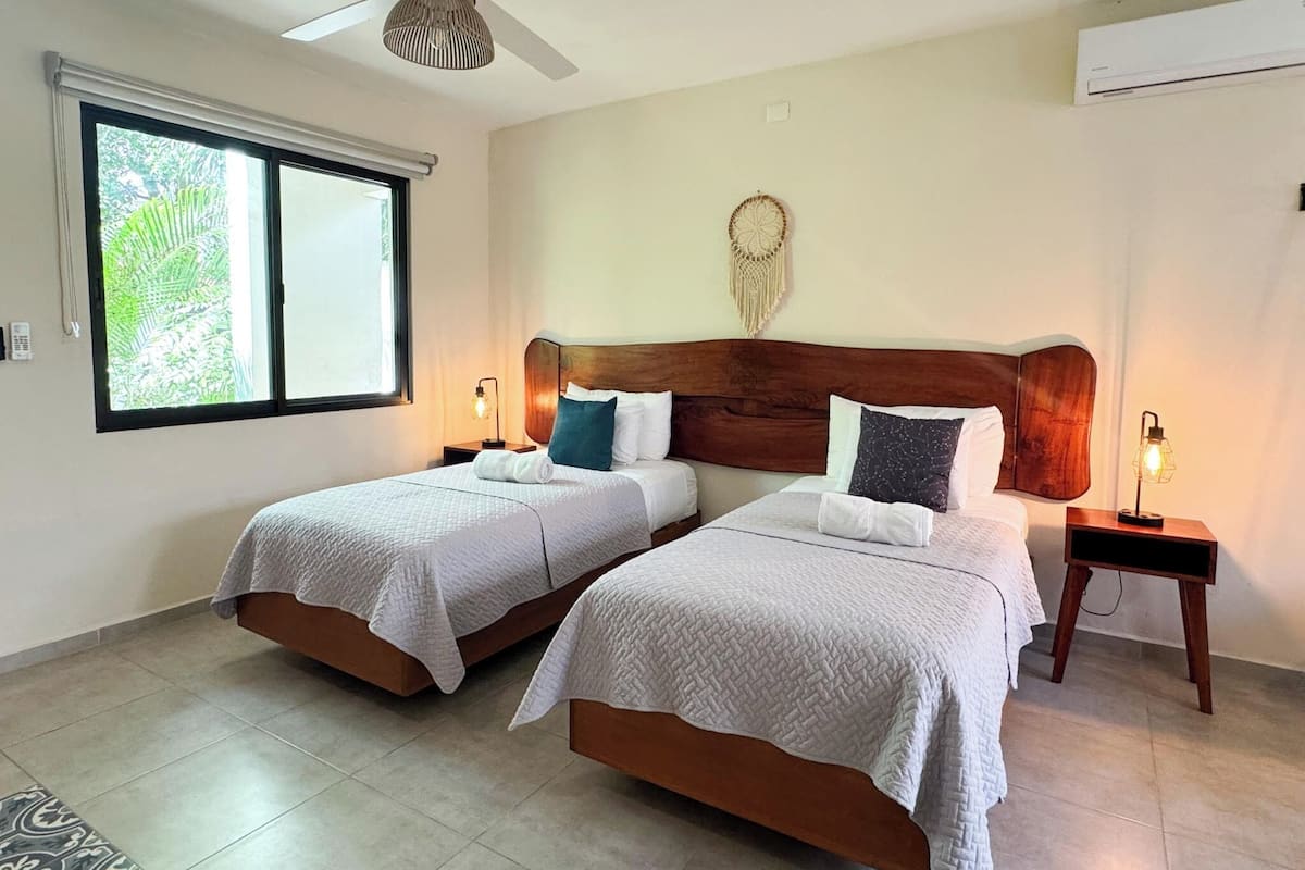 A hotel room with two beds, a ceiling fan, a large window with a view of greenery, and a macrame wall hanging.