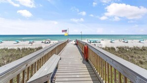 Ein hölzerner Steg führt zu einem Strand mit Sonnenschirmen und einer Flagge.