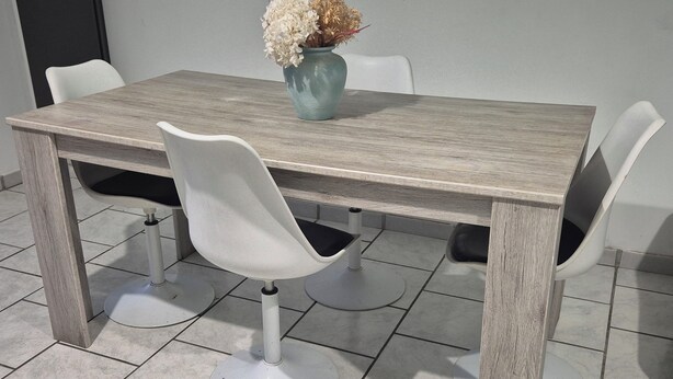 Een rechthoekige tafel met een licht houten oppervlak en vier witte stoelen met zwarte kussens. Op de tafel staat een vaas met gedroogde bloemen.