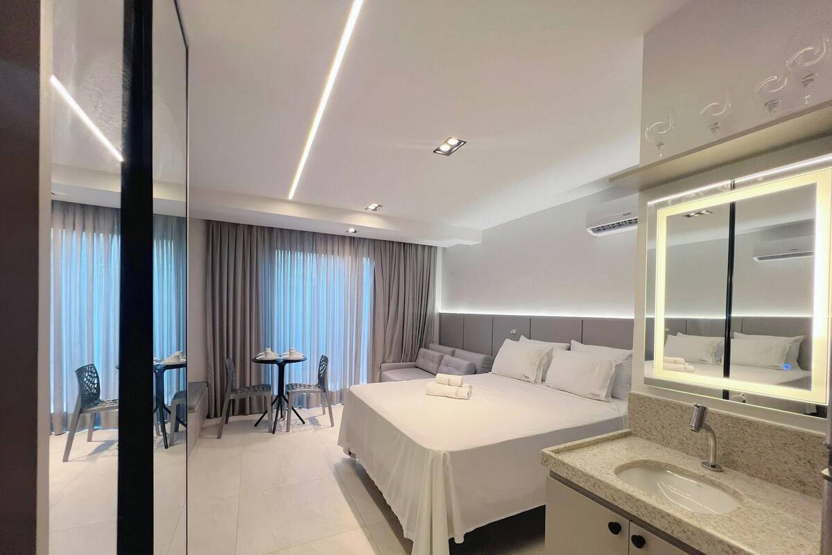 A hotel room with a bed, a bathroom with a mirror and sink, a dining area with chairs and a table, and a view of the outside through large windows.