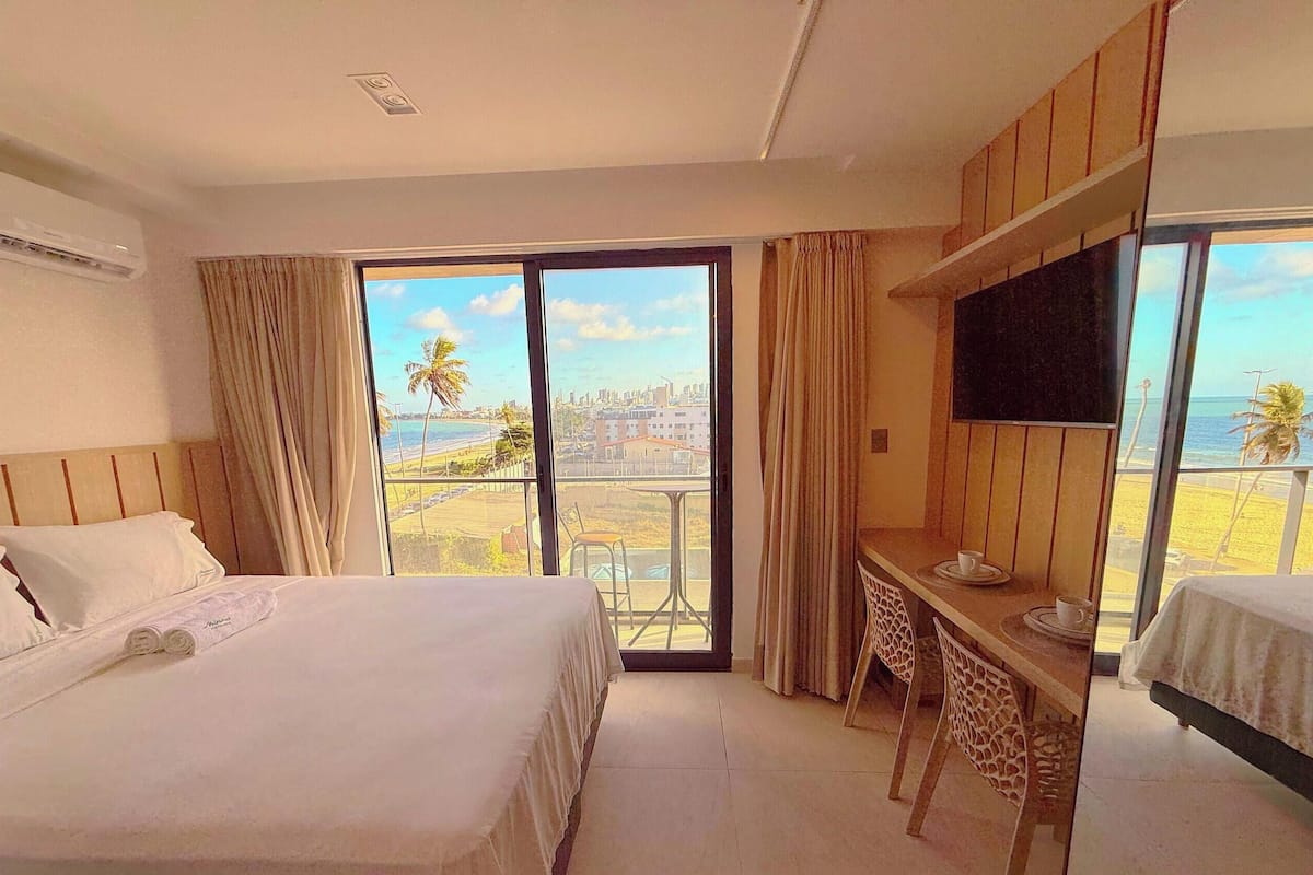 A hotel room with a bed, a desk, a TV, and a balcony with a view of the beach and city.