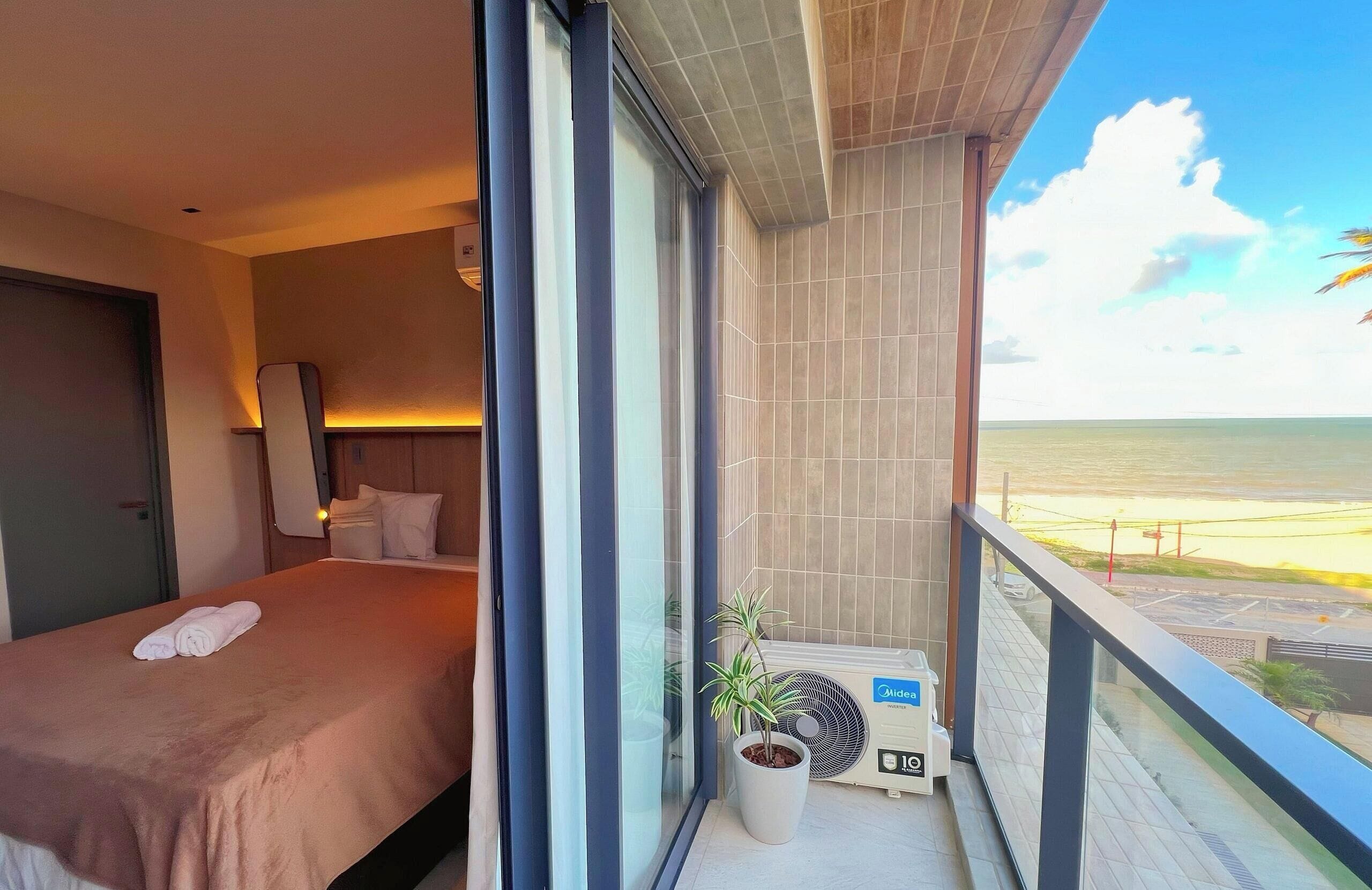 A hotel room with a bed, a potted plant, an air conditioning unit, and a balcony with a view of the beach.