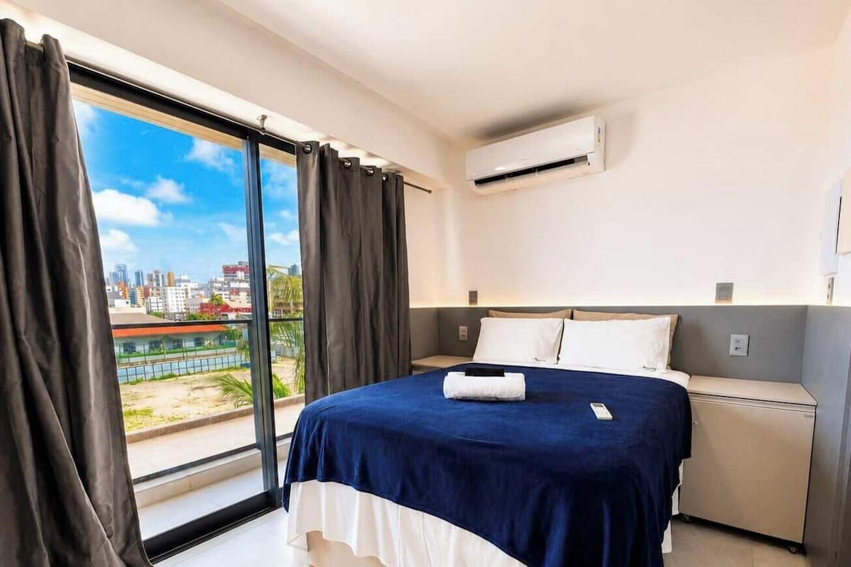 A hotel room with a bed, a bedside table, an air conditioning unit, and a view of a cityscape through large windows.