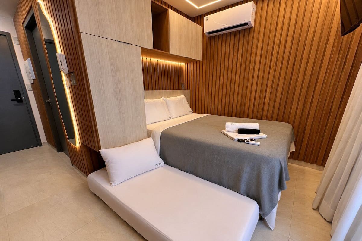 A hotel room with a bed, a bedside table, a wall-mounted air conditioner, and a wooden panel wall.