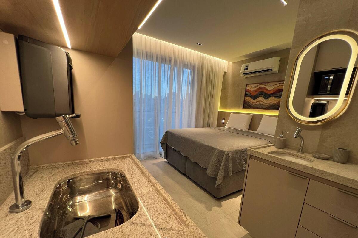A modern hotel room with a built-in sink, a bed with a grey blanket, a bedside table, a mirror, and a large window with curtains.