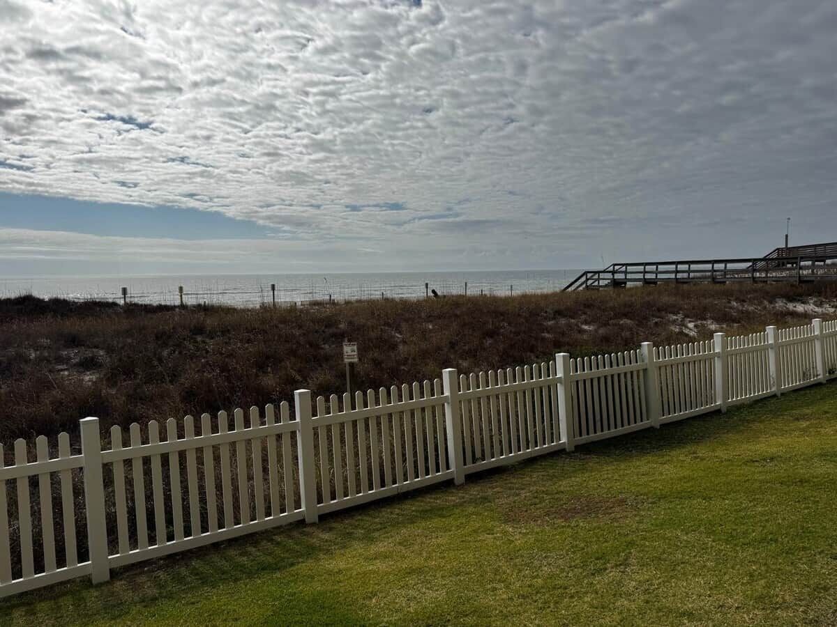 A white picket fence along a grassy area with a view of the beach and ocean.