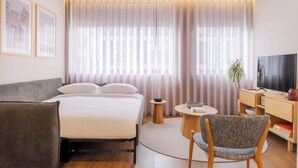 Een hotelkamer met een bed, een bureau, een stoel, een televisie en een groot raam met gordijnen.