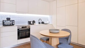 Een moderne keuken met witte kasten, een magnetron, een koffiezetapparaat en een ronde houten tafel met twee stoelen.