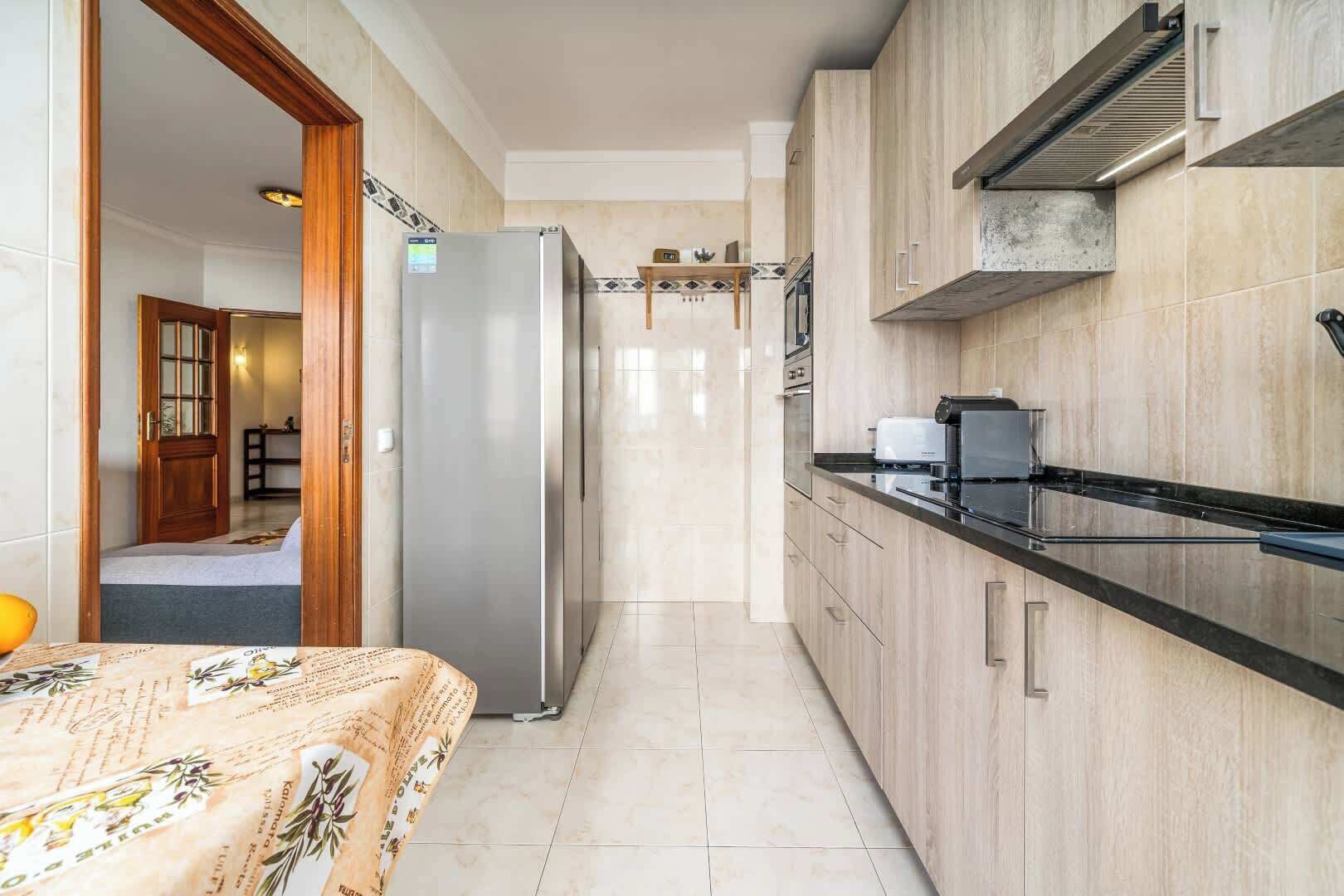 A narrow kitchen with a refrigerator, a stove, and a microwave. There is a wooden door leading to another room.