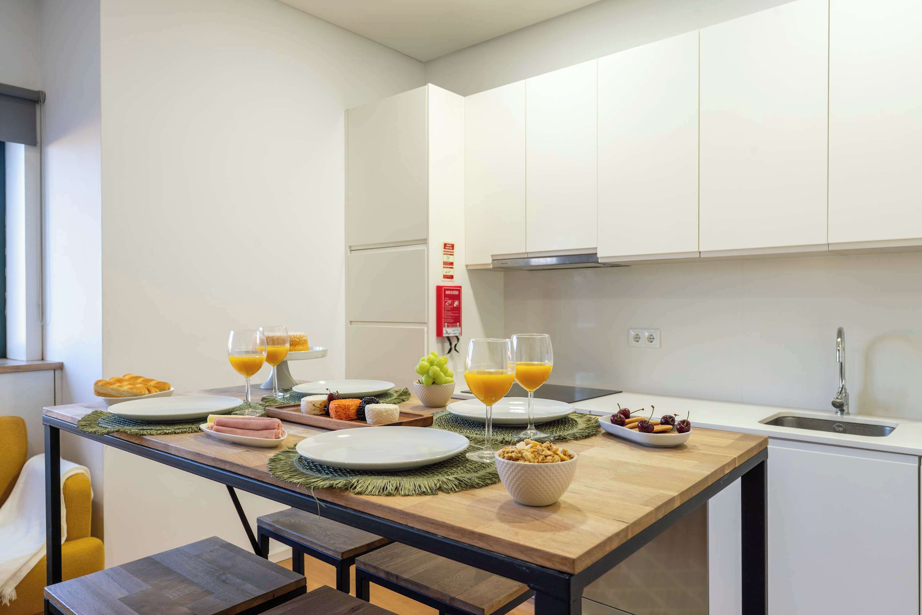 A modern kitchen with white cabinets, a wooden dining table set with plates, glasses, and fruits, and a visible fire extinguisher.