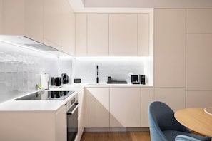 Eine moderne Küche mit weißen Schränken, einer schwarzen Induktionskochplatte, einem eingebauten Backofen und einem blauen Stuhl an einem Holztisch.