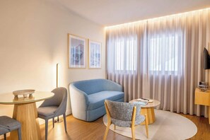 Ein modernes Hotelzimmer mit einem runden Glastisch, einem blauen Sofa, einem grauen Sessel und einem Fernseher auf einem Holzboden.