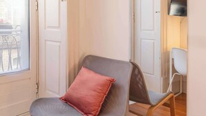 A room with a wooden floor, a grey chair with a red cushion, a white door, and a mirror reflecting a TV and a chair. - Porto Lounge Hostel - Cozy Bright Room w Balcony (Porto)