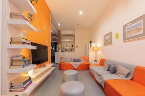 Ein modernes Wohnzimmer mit orangefarbenen Sofas, einem Flachbildfernseher, einem Bücherregal voller Bücher und einer Küche im Hintergrund.