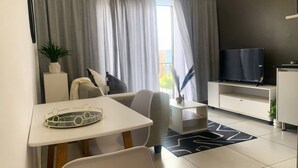 A modern living room with a dining area, a flat-screen TV, and a large window with curtains. - Apartment with balcony in Ferndale, Randburg. Stylish, secure and comfortable. (Randburg)