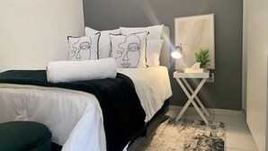 A bedroom with a bed, a bedside table with a lamp, a wall-mounted mirror, and a framed artwork. - Apartment with balcony in Ferndale, Randburg. Stylish, secure and comfortable. (Randburg)
