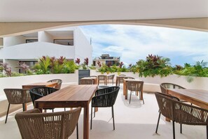 An outdoor patio area with wooden tables and wicker chairs, surrounded by lush greenery and a view of a modern building. - Modern Tulum Studio - Pool + Rooftop + Gym (Tulum)