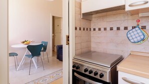 Una cocina con estufa a gas, horno y armarios blancos. Hay una mesa de comedor con sillas y un frutero.