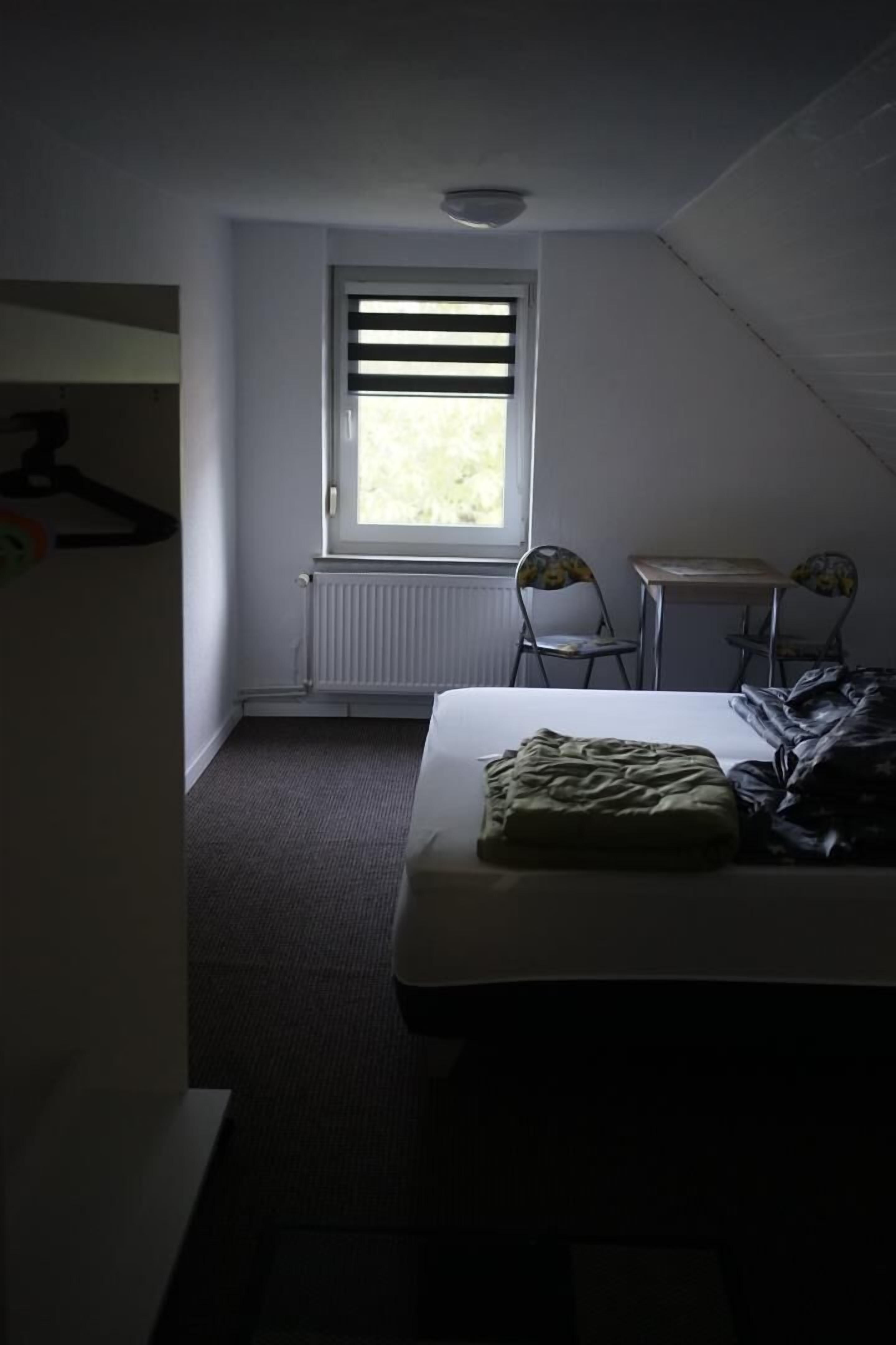 Ein Schlafzimmer mit einem Bett, einem Fenster mit Jalousien, einem Heizkörper und einem kleinen Tisch mit zwei Stühlen.