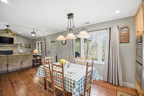 A dining area with a table set for four, a stone fireplace, and a television mounted on the wall. - Coweeta Rapids: Your Elevated Creekside Escape (Otto)