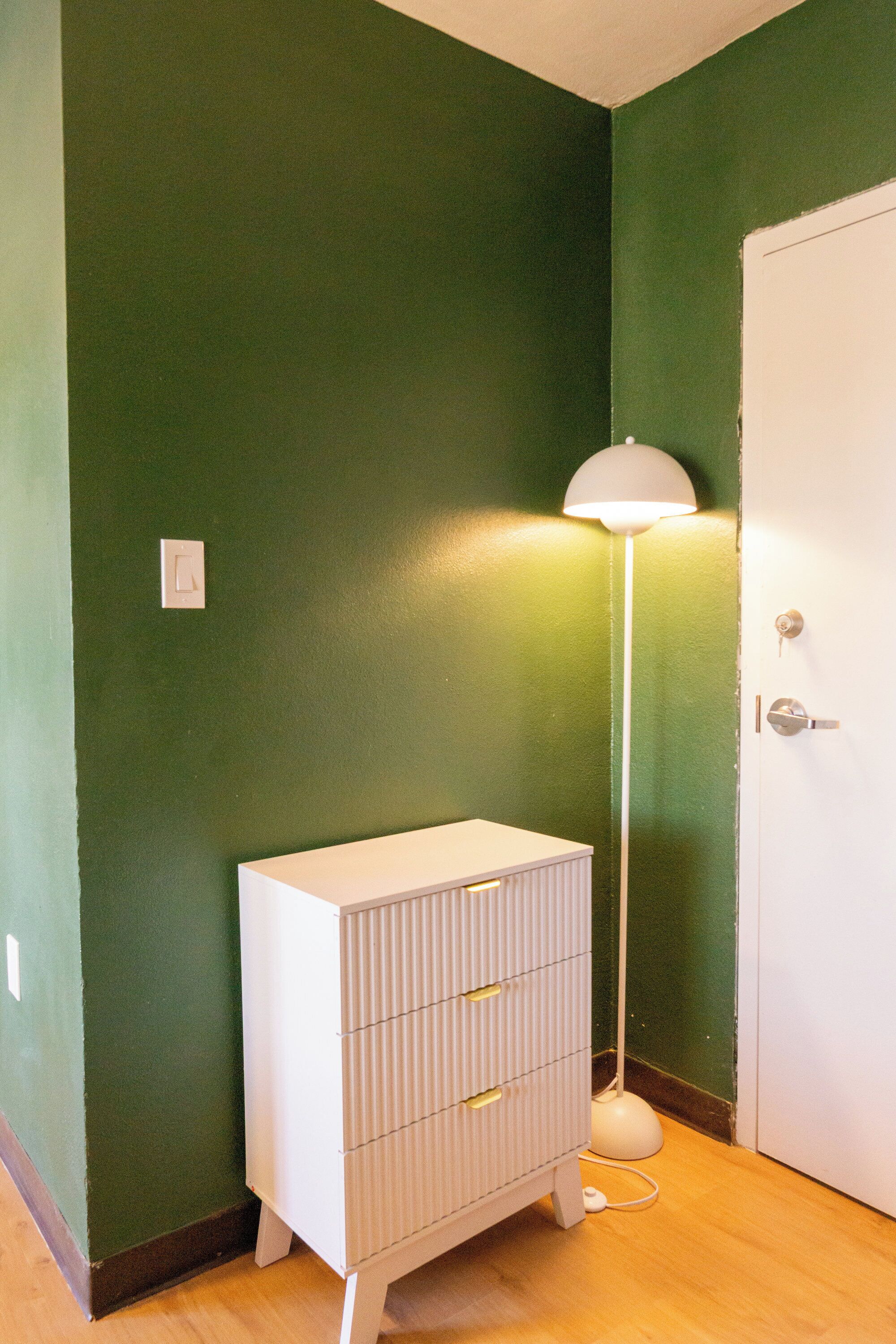 A room with a green wall, a white door, a white dresser with drawers, and a floor lamp.