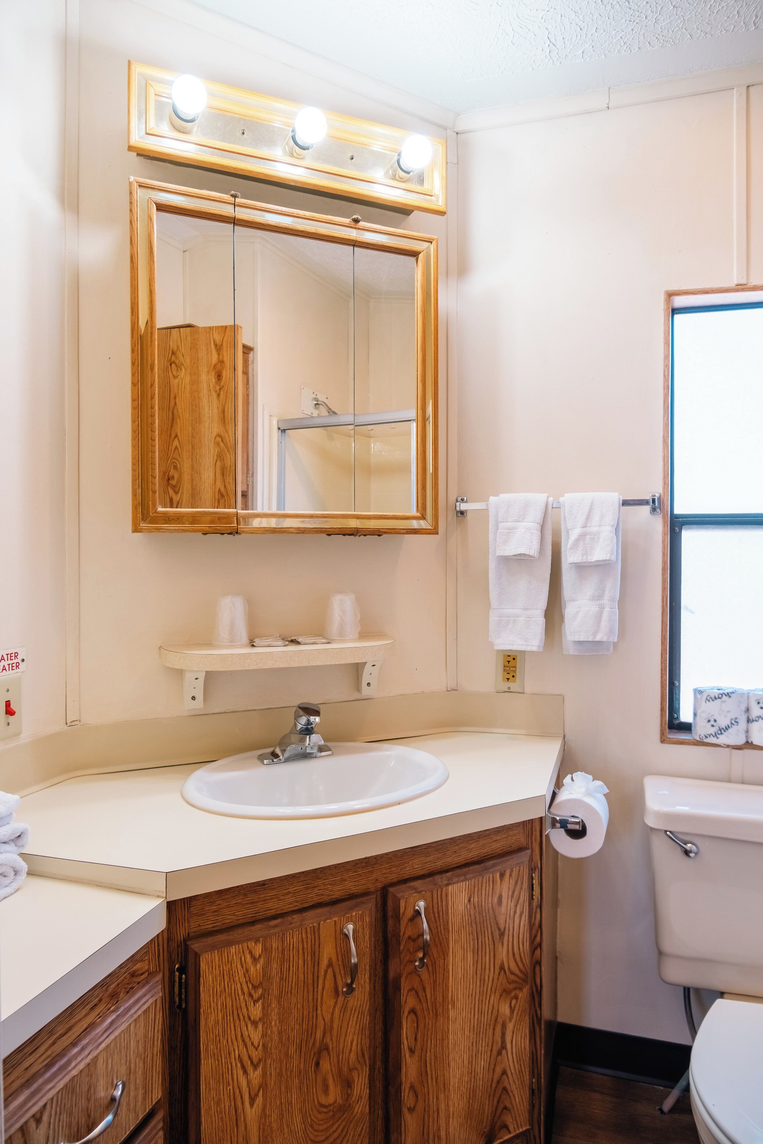 A bathroom with a wooden vanity, a sink, a mirror with lights, a toilet, and a window with a view.