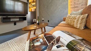 A modern living room with a flat-screen TV, a wooden coffee table with wine glasses and a bottle, a sofa with patterned cushions, and a magazine on the floor.