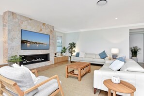 A modern living room with a stone fireplace, a flat-screen TV mounted on the wall, a white sofa, a wooden coffee table, and a comfortable armchair. - Tilligerry Beach Retreat (Taylors Beach)