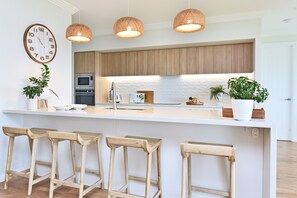 A modern kitchen with wooden cabinets, a white countertop, a clock, hanging lights, and bar stools. - Tilligerry Beach Retreat (Taylors Beach)