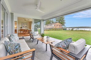 A modern living room with a large glass door leading to a dining area, a view of the sea, and a well-maintained lawn. - Tilligerry Beach Retreat (Taylors Beach)