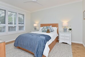 A bedroom with a bed, two bedside tables, a nightstand, a window with blinds, and a ceiling fan. - Tilligerry Beach Retreat (Taylors Beach)