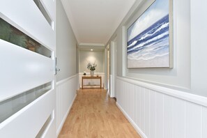 A hallway with white wainscoting, a wooden floor, a painting of a beach scene, and a small table with a vase of flowers. - Tilligerry Beach Retreat (Taylors Beach)