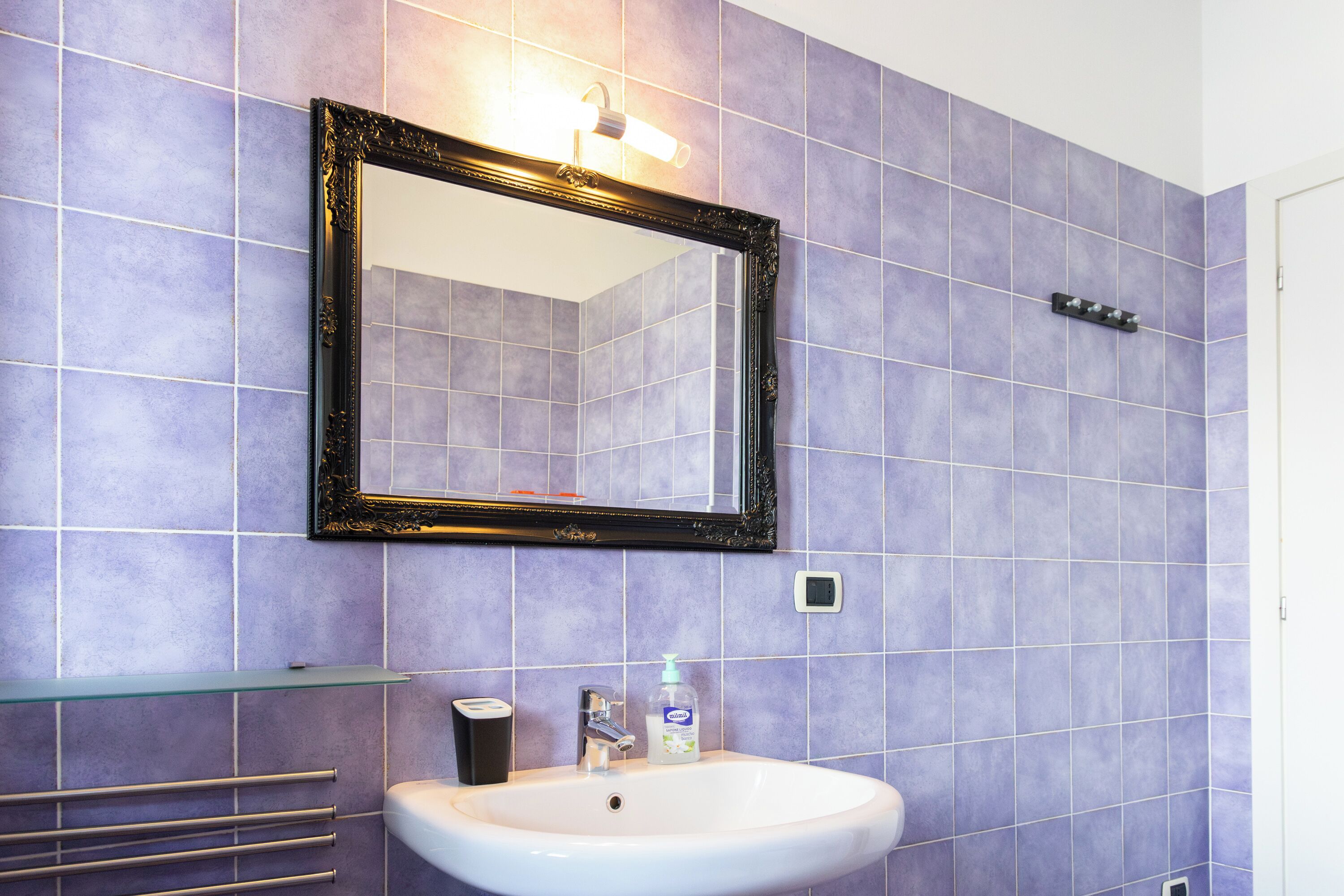 Une salle de bain avec des carreaux violets, un lavabo blanc, un miroir à cadre noir et un distributeur de savon.