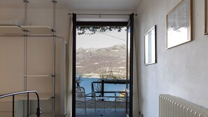 Una habitación con cama, silla, mesa, radiador y vistas a montañas por una puerta.