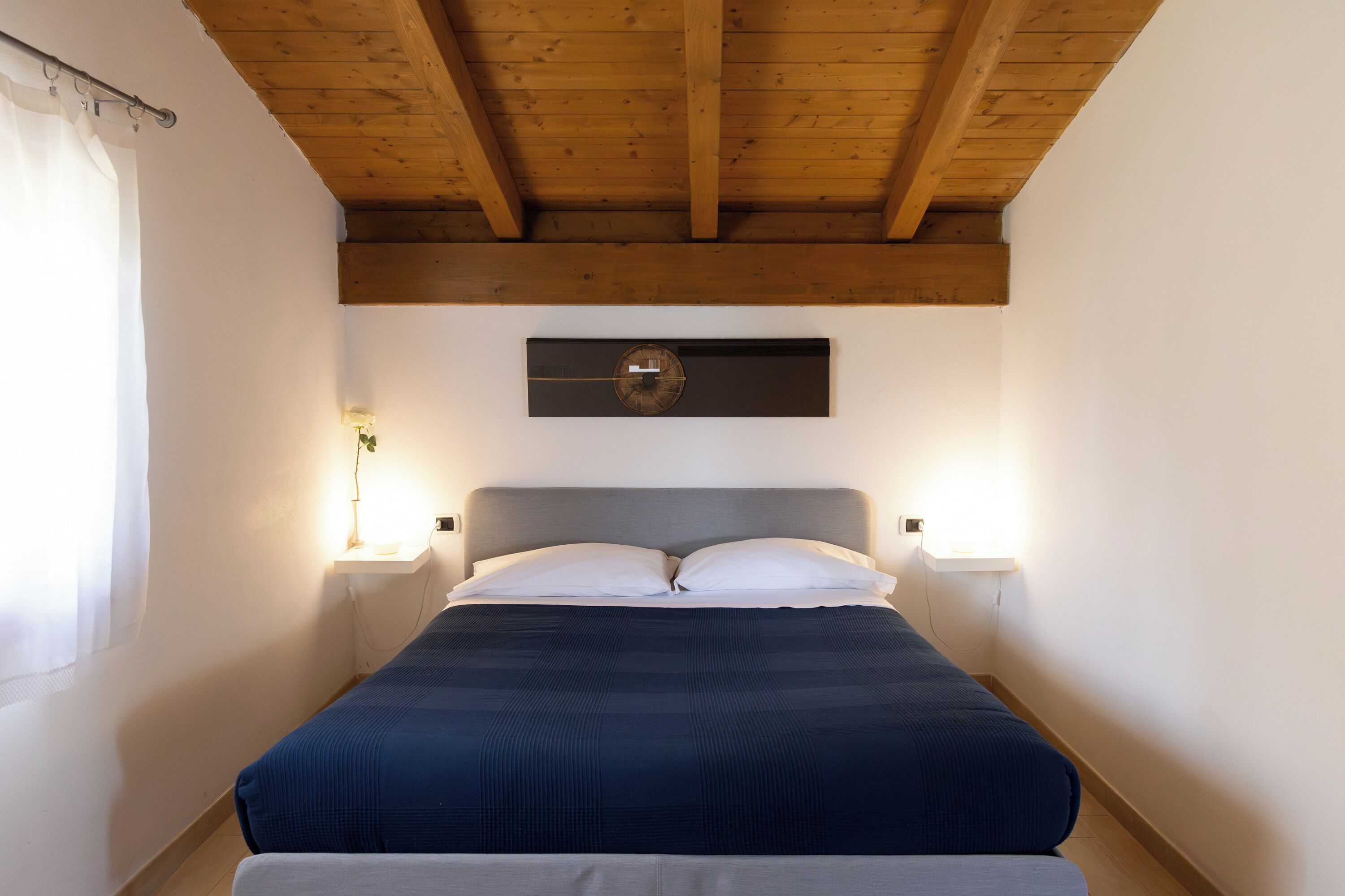 A bedroom with a bed, wooden ceiling beams, a wall clock, bedside lamps, and a window with curtains.