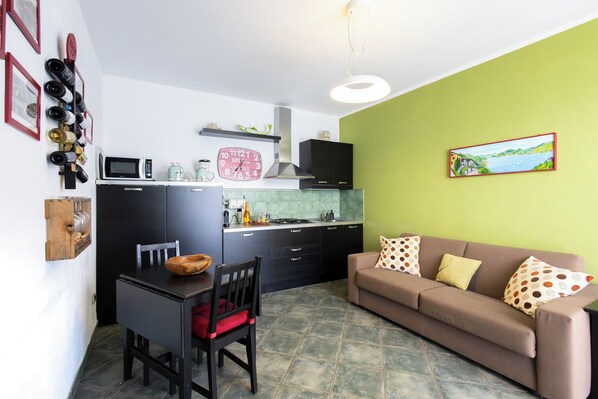 Ein kompakter Wohnraum mit Küchenzeile, einer Essecke mit Tisch und Stühlen, einer Couch mit gemusterten Kissen und einem an der Wand befestigten Weinkühlschrank.