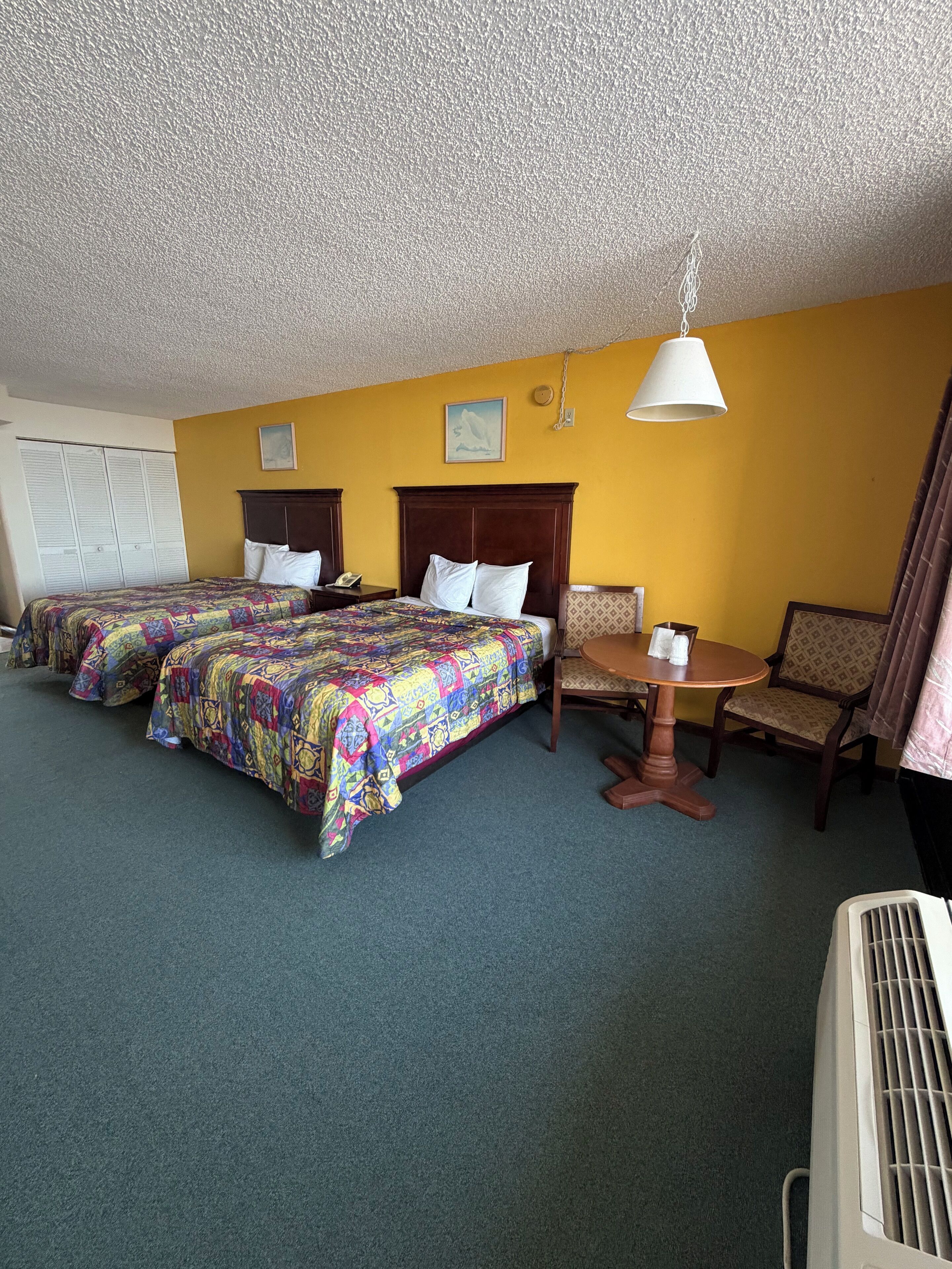 A hotel room with two beds, a small table, chairs, a ceiling fan, and a window with curtains.