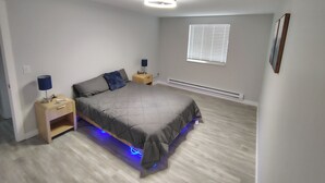 Quarto com cama, mesas de cabeceira, janela com persianas, luminária de teto e aquecedor fixado na parede.
