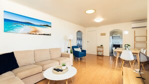 Ett vardagsrum med en beige soffa, en blå fåtölj, ett runt soffbord och en tavla som föreställer en strandvy.