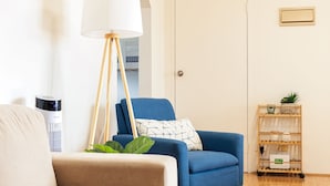 Ett vardagsrum med en blå fåtölj, en beige soffa, en golvlampa, en värmare och en trähylla med växter.