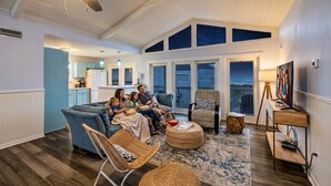 A spacious living room with a high ceiling, a ceiling fan, a large window with a view, and a cozy seating area. - Coastal/Cozy- Steps to the sand and sea (Jamaica Beach)