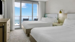 Une chambre d’hôtel avec deux lits, une table de chevet avec une lampe et une vue sur la plage grâce à de grandes fenêtres.
