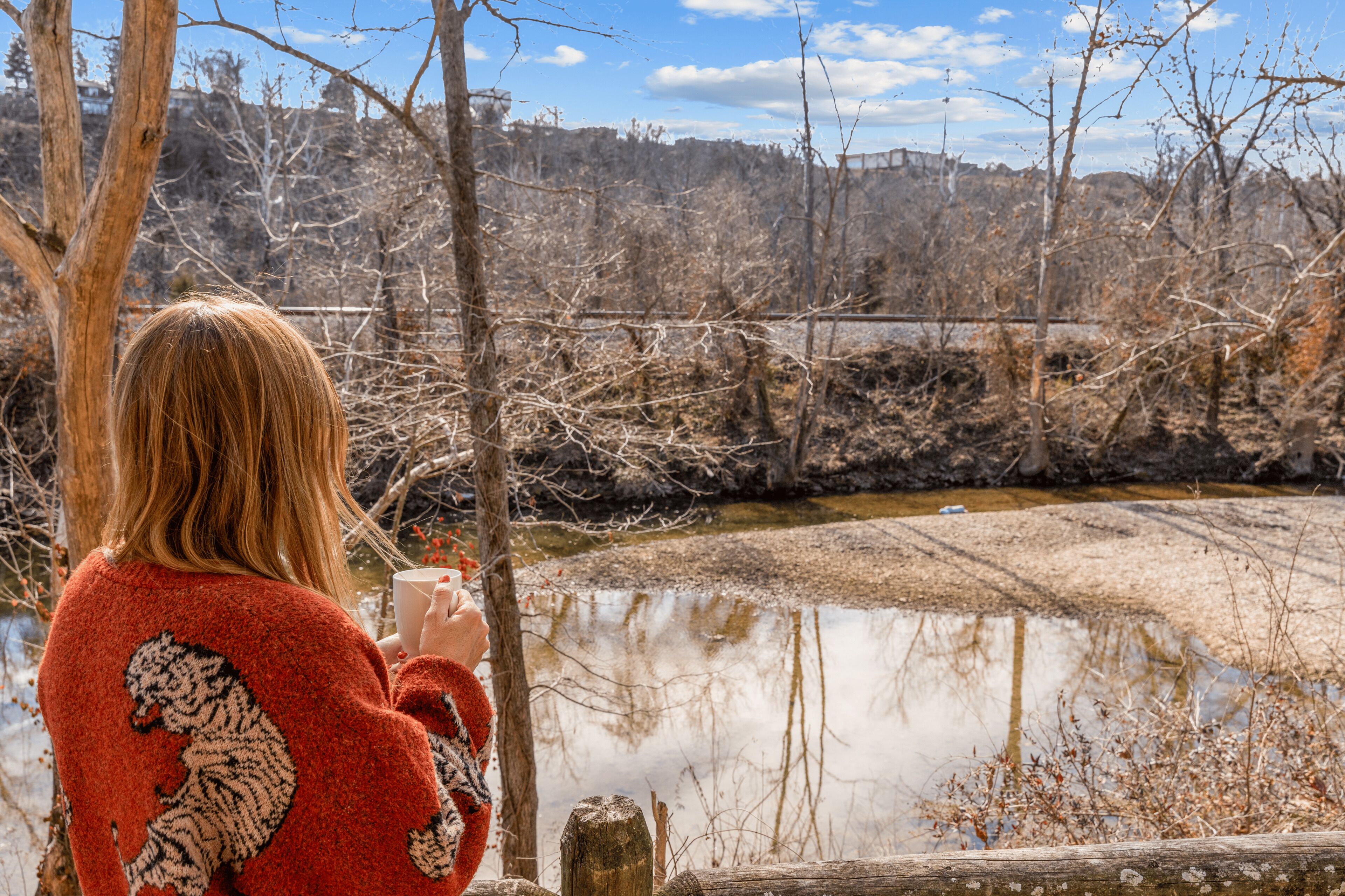 A person in a red sweater with a lion design, standing by a body of water with trees and a fence in the background.