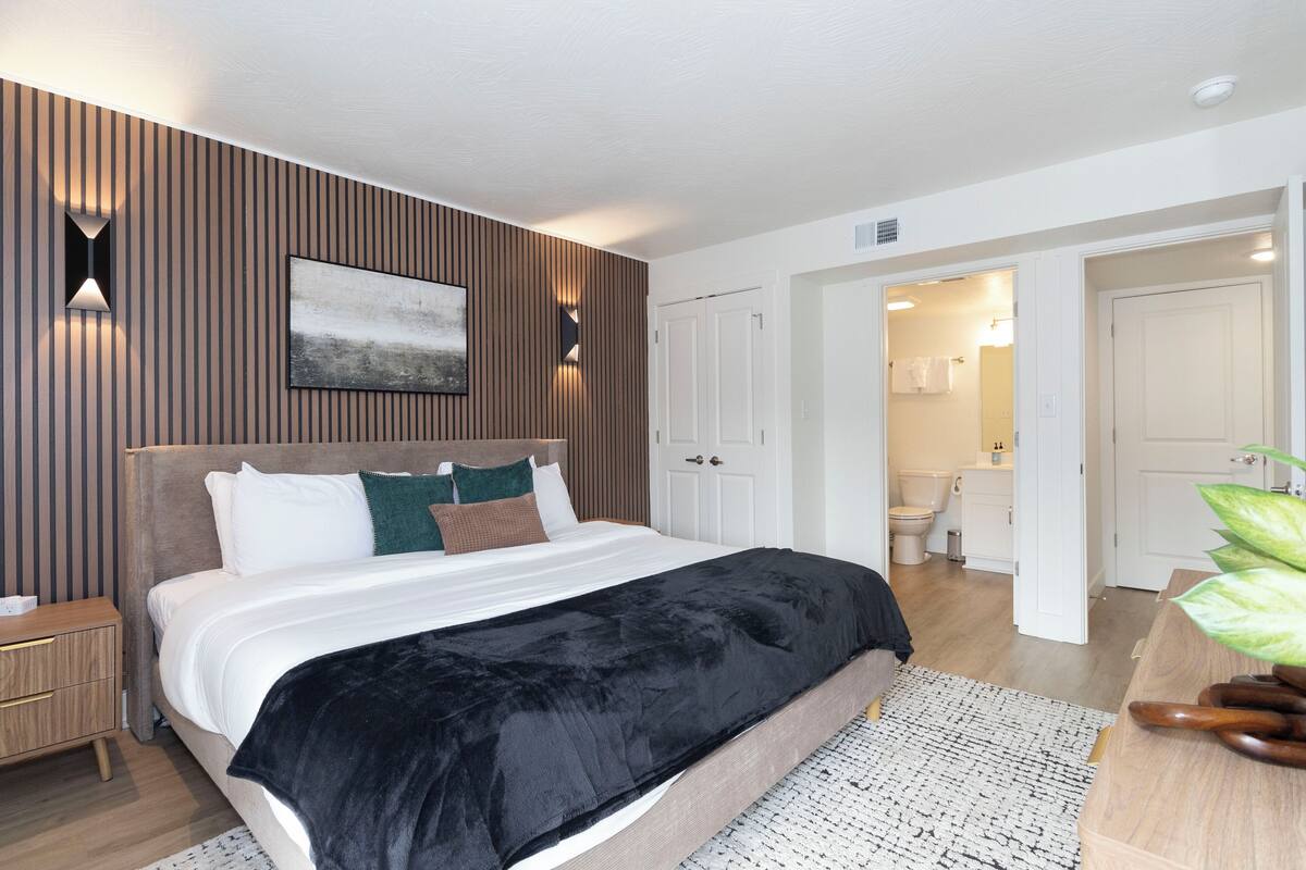 A modern bedroom with a large bed, a bedside table, a wall-mounted light, and a door leading to a bathroom.